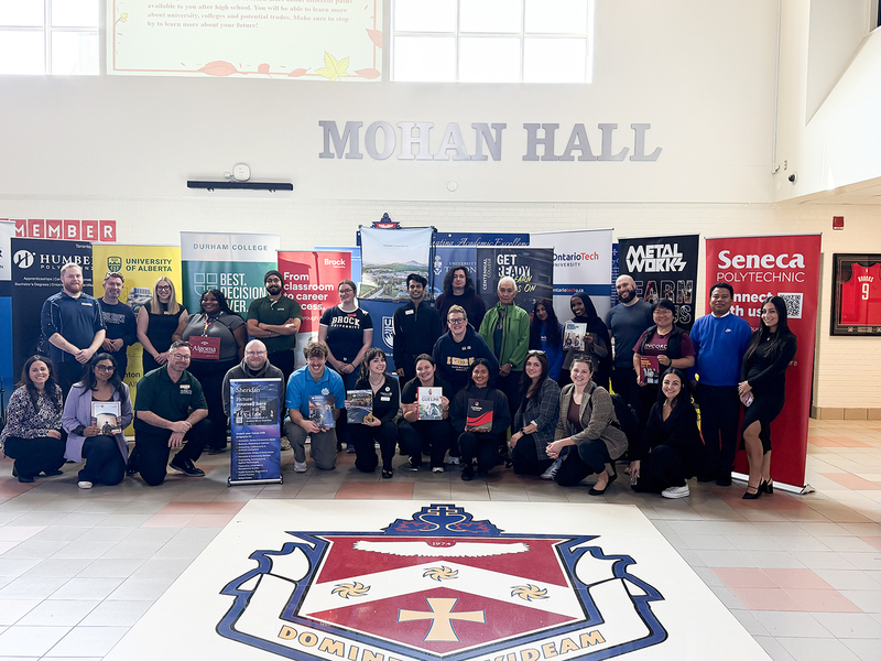 Group photo of presenters at the fair with their organization's banners behind them, posing in the FHC school building, with the FHC school crest emblazoned on the floor in front of them