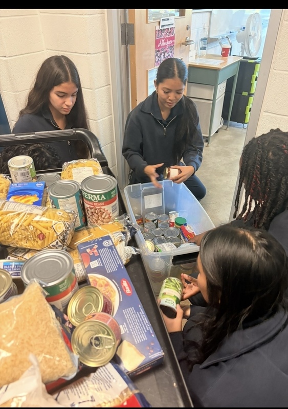 Two people sit at a table covered with food items and canned goods, sorting products while one checks a phone; a blue-lidded container and many packages are spread across the tabletop in an indoor room.