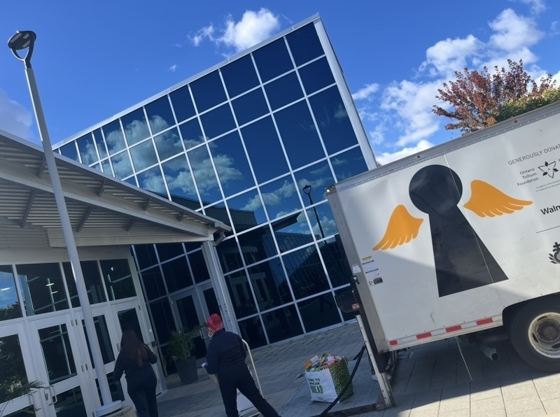 A modern building with large blue glass panels under a bright sky, with two people walking near the entrance and a parked truck beside a wall featuring orange graphic shapes.