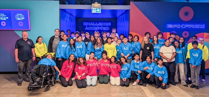 Group photo of Redmond students posing in blue, red and yellow team shirts along with Redmond staff in front of a neon "Challenge Accepted" sign at BRKFREE
