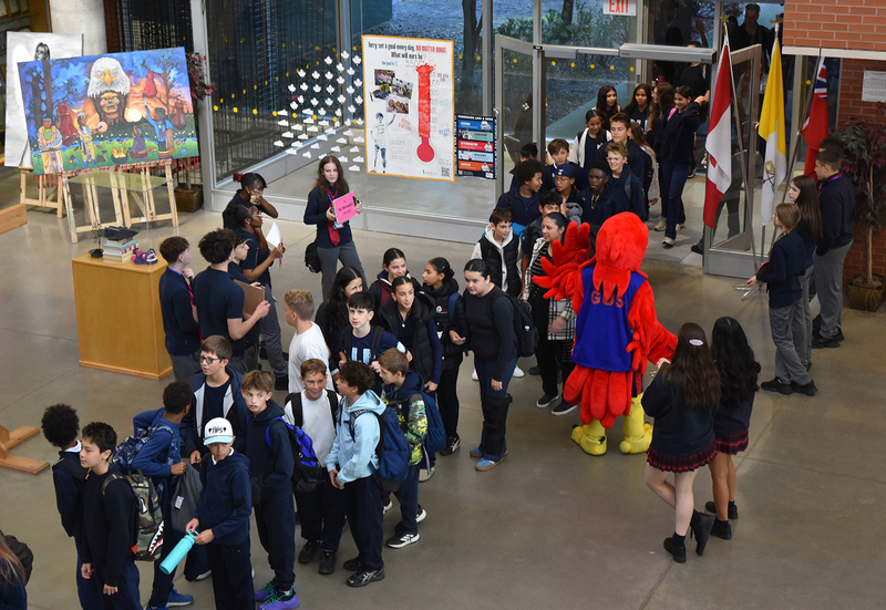Photo of elementary students waiting in line to get into the festival, while Redmond students hold signs to welcome and guide them, along with a mascot in red bird costume