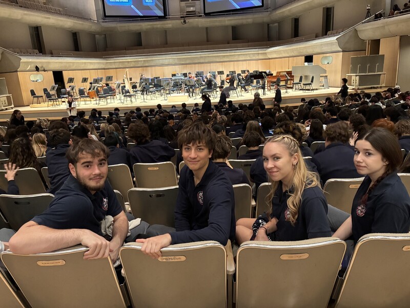 Students in a lecture hall sit in rows of gray seats, smiling and posing for the camera while others sit in the background, with a stage and orchestra instruments set up at the far end.