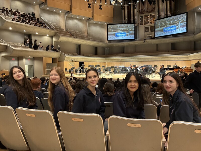 Students in a lecture hall sit in rows of gray seats, smiling and posing for the camera while others sit in the background, with a stage and orchestra instruments set up at the far end.