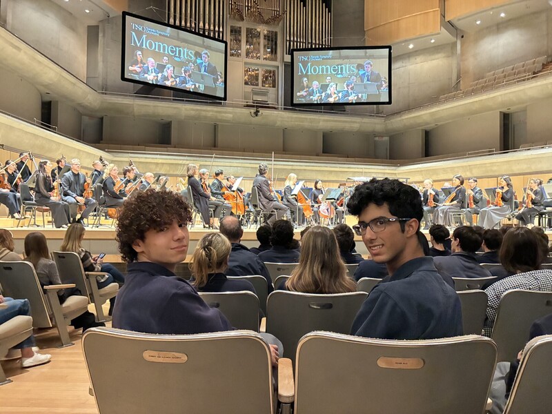 Students in a lecture hall sit in rows of gray seats, smiling and posing for the camera while others sit in the background, with a stage and orchestra instruments set up at the far end.