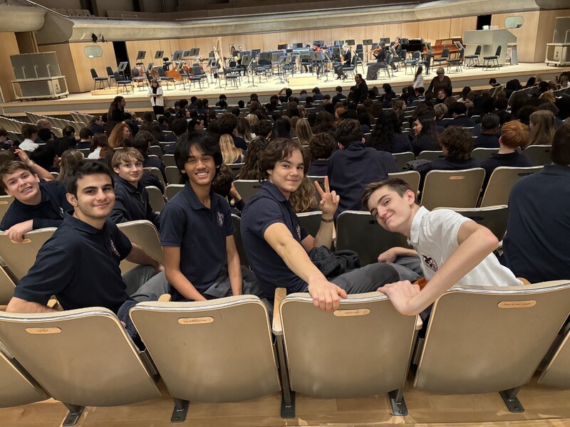 Students in a lecture hall sit in rows of gray seats, smiling and posing for the camera while others sit in the background, with a stage and orchestra instruments set up at the far end.