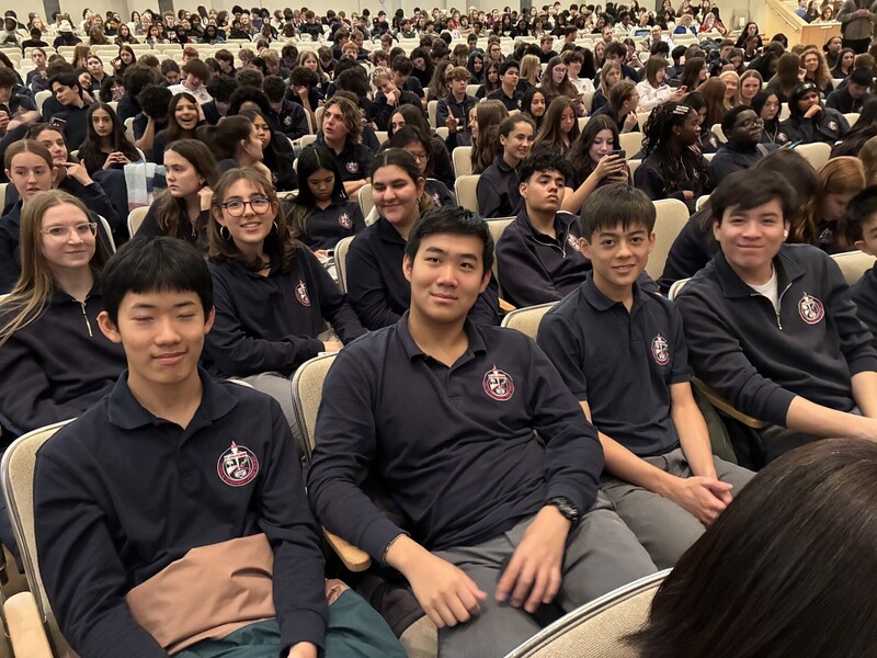 Students in a lecture hall sit in rows of gray seats, smiling and posing for the camera while others sit in the background, with a stage and orchestra instruments set up at the far end.