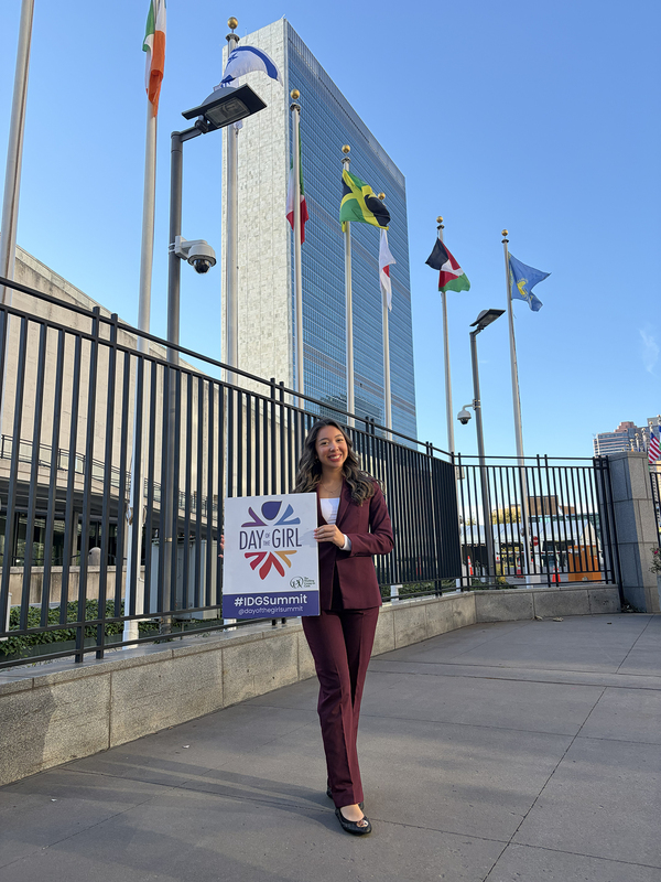 Photo of Sofia posing with a flyer avertising the summit, with the flags of various countries flying behind her