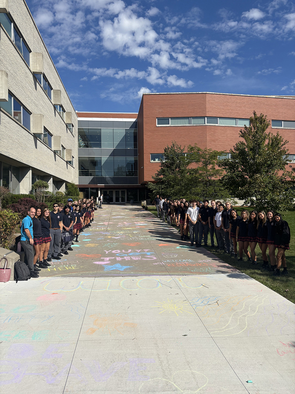 Photo of FJR students and staff in front of the school building with the welcoming chalk messages scrawled on the pavement