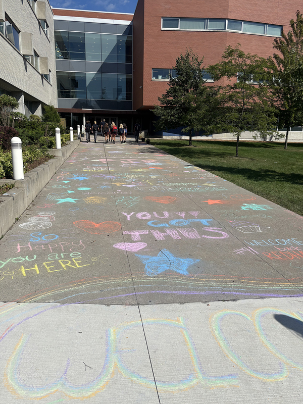 Photo of FJR students and staff walking into the school building with the welcoming chalk messages scrawled on the pavement behind them