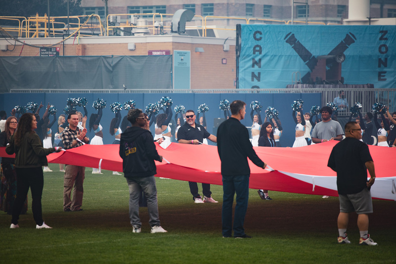 Photo of Coach Vescio and students on the field carrying along a huge Canada flag