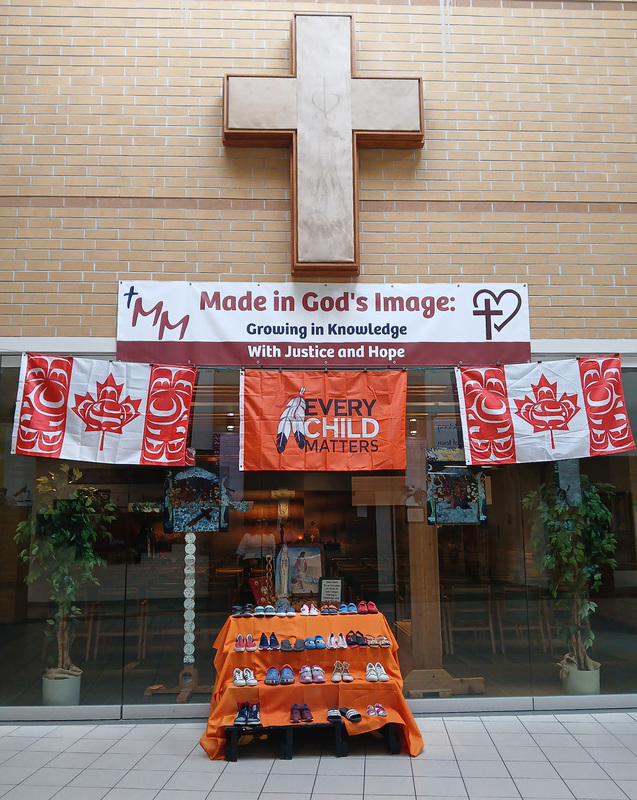 Photo of the shoe display with the Every Child matters flag above it and two Canada flags with indigenous graphics flanking it, a banner saying "Made in God's image: Growing in Knowledge With Justice and Hope" above, and a cross hanging from the wall above that.