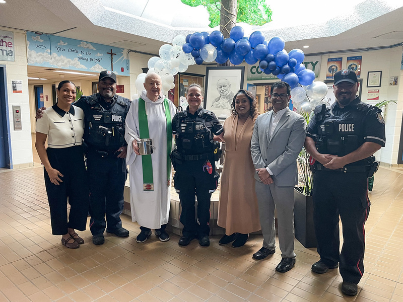 Group photo of the priest, the Chaplain, police officers, Principal Navaratnasingham and Superintendent Peterson in front of a drawing of St. John Paul II