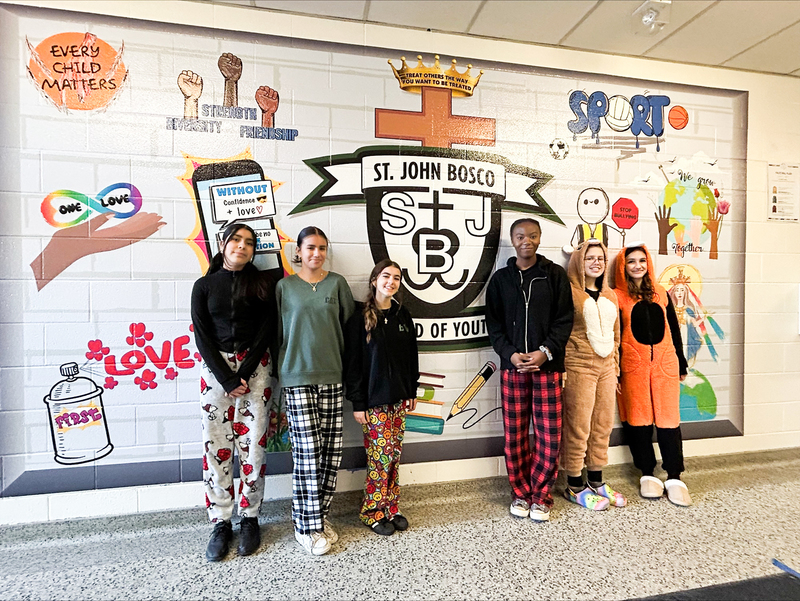 Photo of St. John Bosco students in comfy clothing in front of a school wall with the St. John Bosco crest