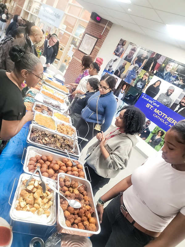 Photo of  students serving food at the celebration while students and staff peruse the food available