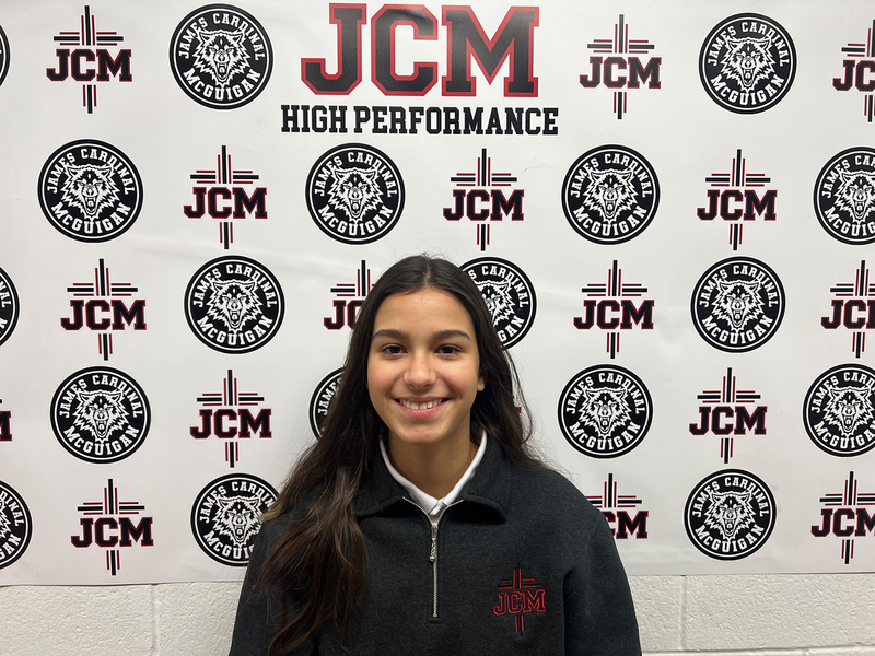 Photo of Cecilia standing in front of a wall papered with the JCM High Performance and Wolfpack logo