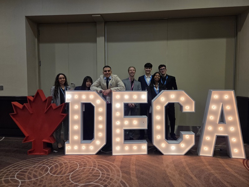 7 students posing dressed up in business attire in front of giant lit up letters that spell "DECA"