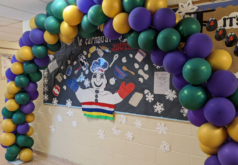 A classroom board decorated with a paper bonhomme and paper snowflakes and other graphics around a balloon arch for Carnaval