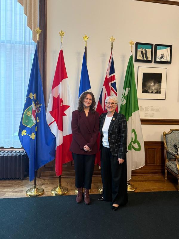 Two women posing in front of flags while dressed in business attire.