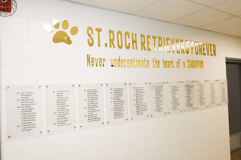 Photo of the gym wall showing a display of St. Roch sports championships and achievements with the text: St. Roch Retrievers Forever - Never underestimate the heart of a CHAMPION