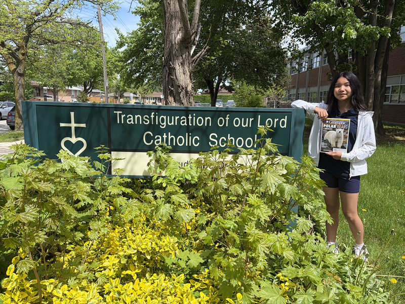 Photo of Sinead holding up a Canadian Geographic magazine next to the Transfiguration of our Lord school sign in front of the school building