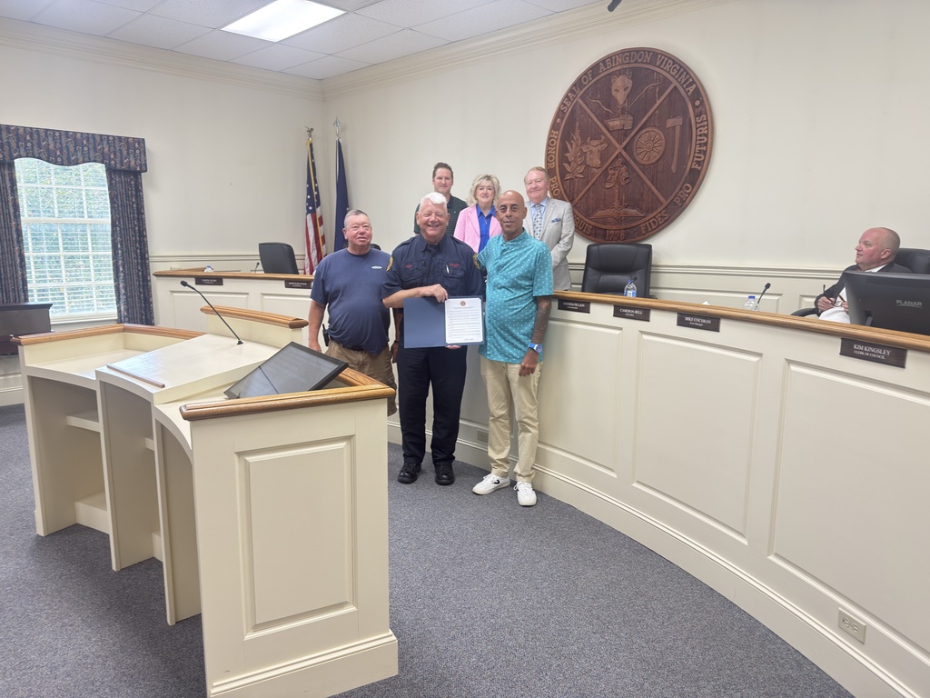Four people stand in a council meeting room, two holding a paper. A podium is in front of them.