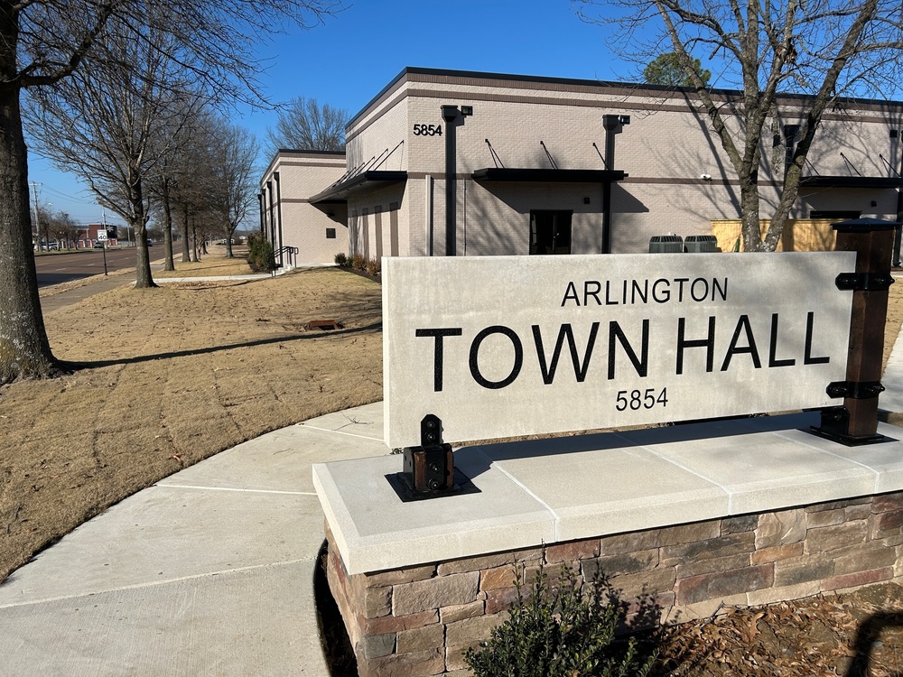 Photo of Arlington Town Hall monument sign, in beige concrete with a stone base. In background is a beige brick building with bronze awnings and details. The sky is bright blue and trees lining the street in front of the building are bare of their leaves.
