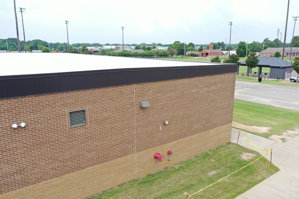 Picture of corner of a two-tone beige brick building with dark bronze flashing and white roof. Grass play fields are in the background, with a brick fire station and other buildings beyond.