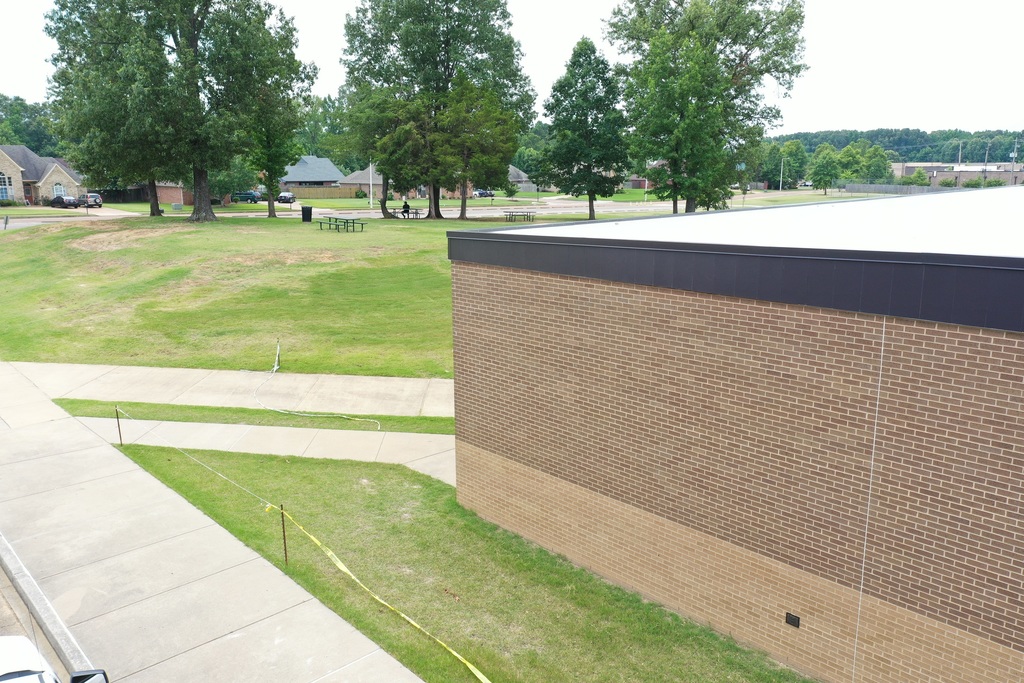 Picture of corner of a two-tone beige brick building with dark bronze flashing and white roof. A grassy hilled park is in the background with several large trees and picnic table