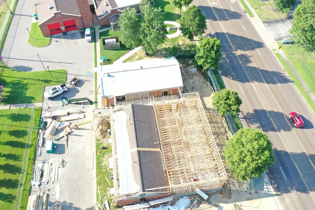 Aerial picture of construction site with an existing building alongside a wood framed expansion, extending toward a tree lined street. A fire station with red roll-up doors is in the background. 