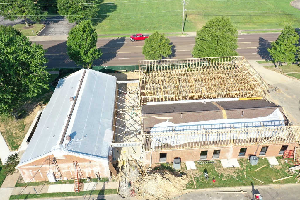 Aerial picture of two brick buildings on a construction site. The existing buildings have white tarps covering the roof and are attached to a large wood framed expansion area, as well as a connection between the two that is being framed in. The building sits on a 5-lane, tree-lined road which is visible in background.
