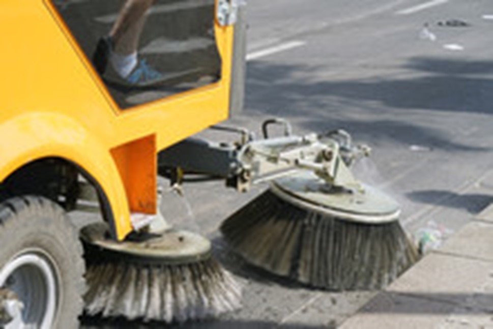 Picture of front of a yellow street sweeper, with round brushes running along the curb on a street.