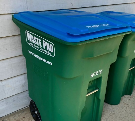 Picture of two large residential trash bins sitting in front of gray, wood siding. Bins are green with blue lids and say Waste Pro on the side