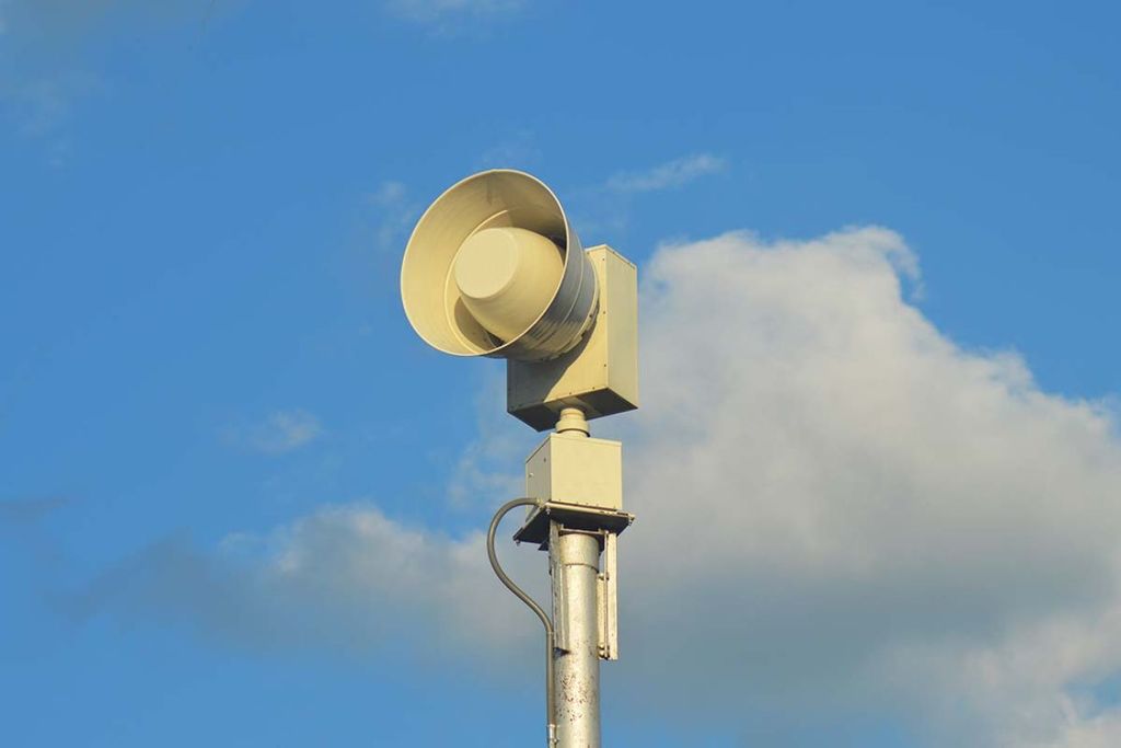 Beige Tornado Siren sitting atop a silver pole with blue sky and white clouds in background