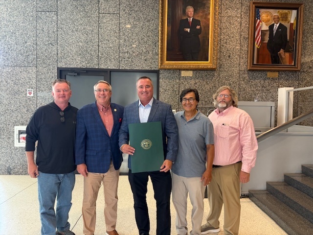 Five men posing with man in center holding official award document. Background is gray granite walls with large portraits of officials on the walls.