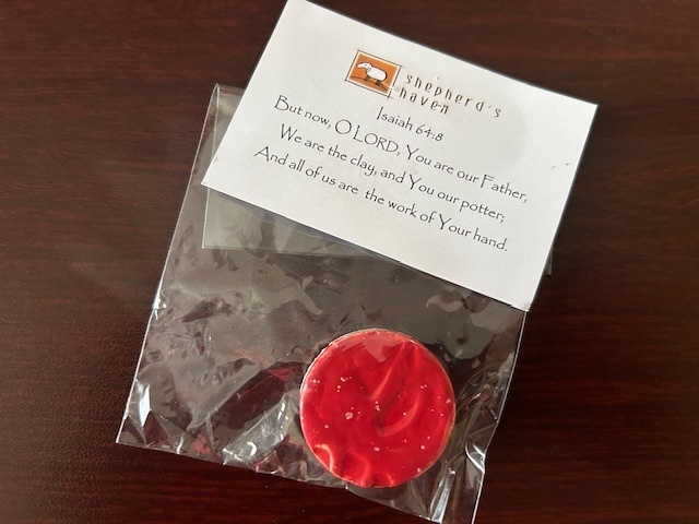 a round, bright red painted ceramic magnet in a plastic bag with a Shepherd's Haven card on the top