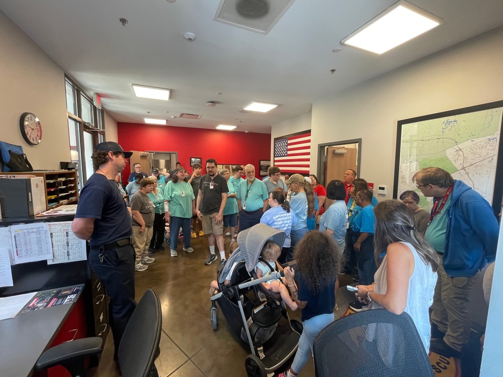 A group of adults gathered in an office area with maps and flags on the wall, listening to uniformed firefighter give a tour
