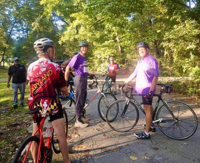 several seniors standing astride bike on a paved trail in a park, two wearing purple arlington shirts