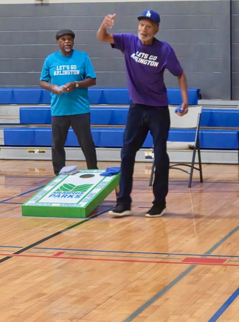 Two seniors men in purple and blue shirts tossing cornhole bags in a gymnasium