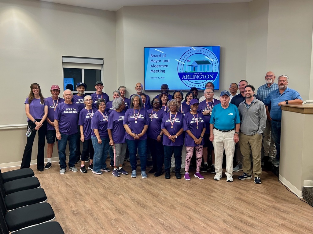 A line of seniors in purple arlington shirts posing in a board room with several elected officials in front of a screen that says Board of Mayor and Aldermen meeting, October 2025