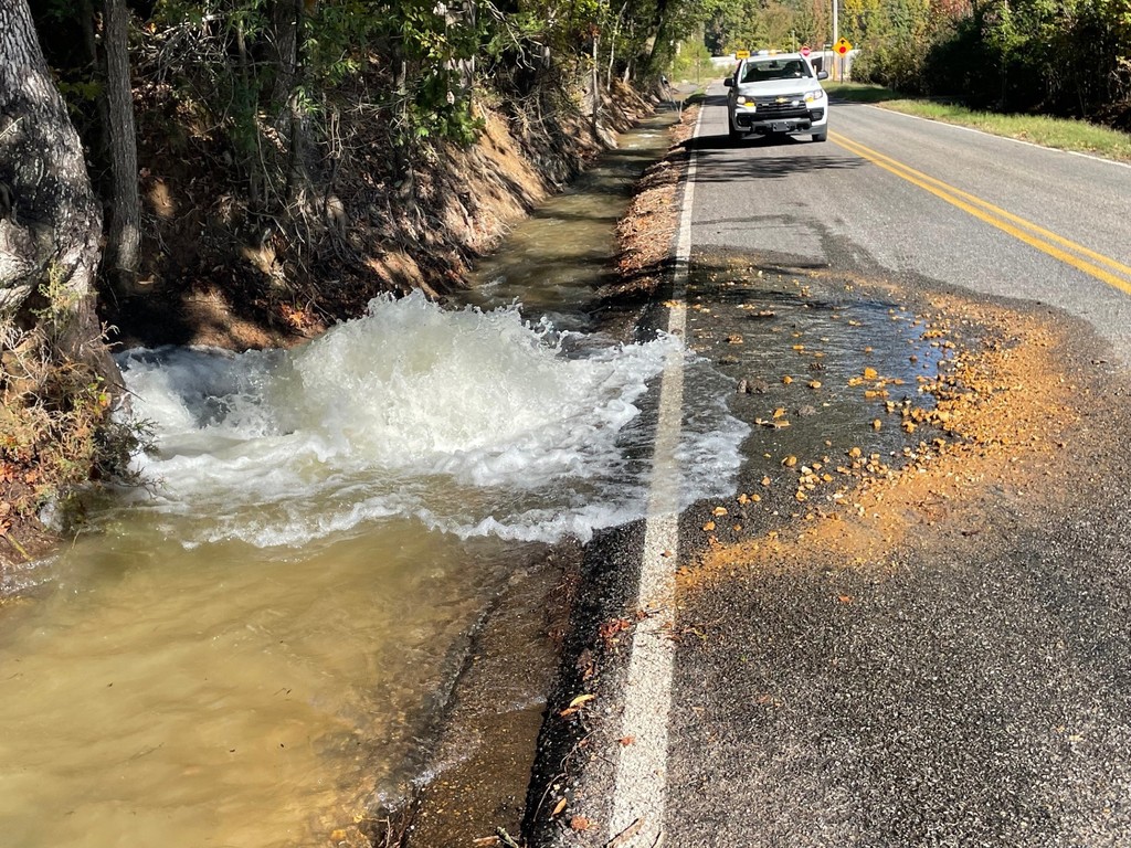 Picture of water bubbling up and filling a ditch along a two-lane road. A white town truck is parked in one lane in background.