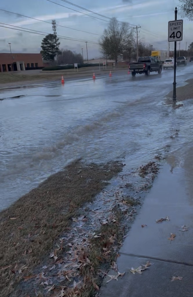 Photo of water pooling in two lanes of a 4-lane road and on sidewalk as traffic passes. Brick Post office can be seen in background