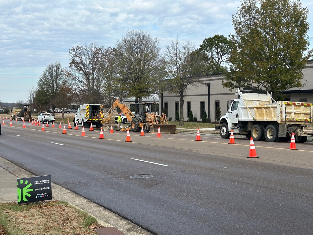 Construction crews, trucks and equipment on a 4-lane road with orange cones directing traffic around a work area. 