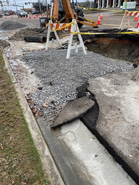 Picture of large hole in a roadway, with caution tape and barriers around it, as well as a backhoe to one side.