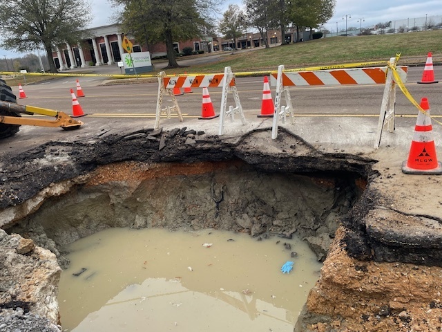 Large hole in roadway that is filled with muddy water. Caution barriers and tape surround the hole.