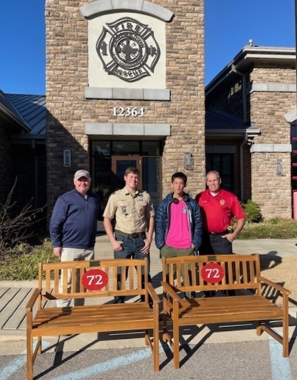 Picture of two scouts, a man in a blue jacket, and a fireman in a red shirt posing behind two wooden benches with red circles and the number 72 on them. all are in front of a stone fire station with blue sky behind