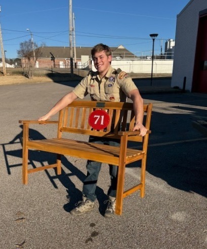 Picture of a teenage scout in uniform and carrying a wooden bench with red circle and number 71 on it. 