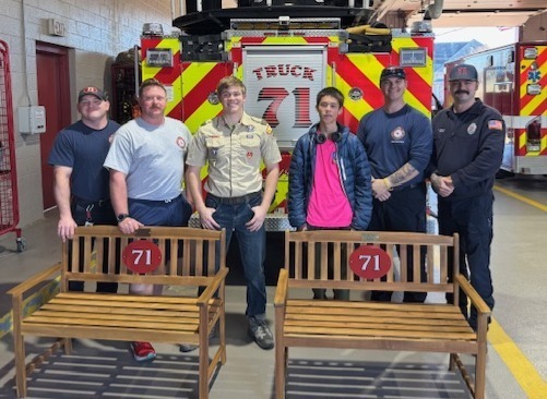Picture of four firemen and two eagle scouts posing behind two wooden benches with red circles and the number 71 on them, all in front of Fire Truck 71 .