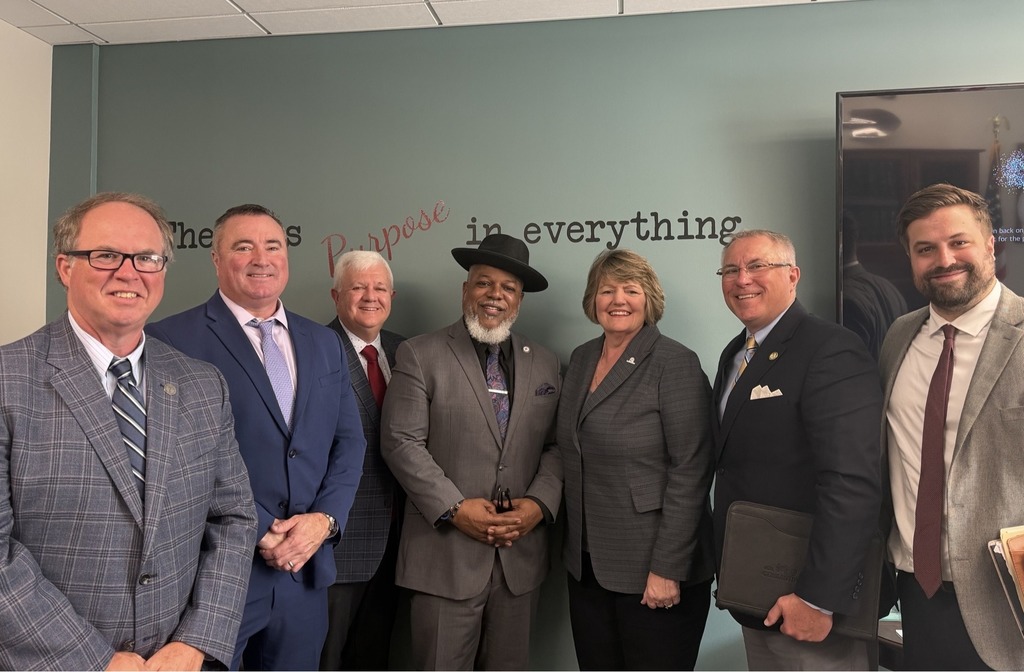 7 political officials in suits posing in front of a light blue wall with a phrase on it, partially covered by the officials 