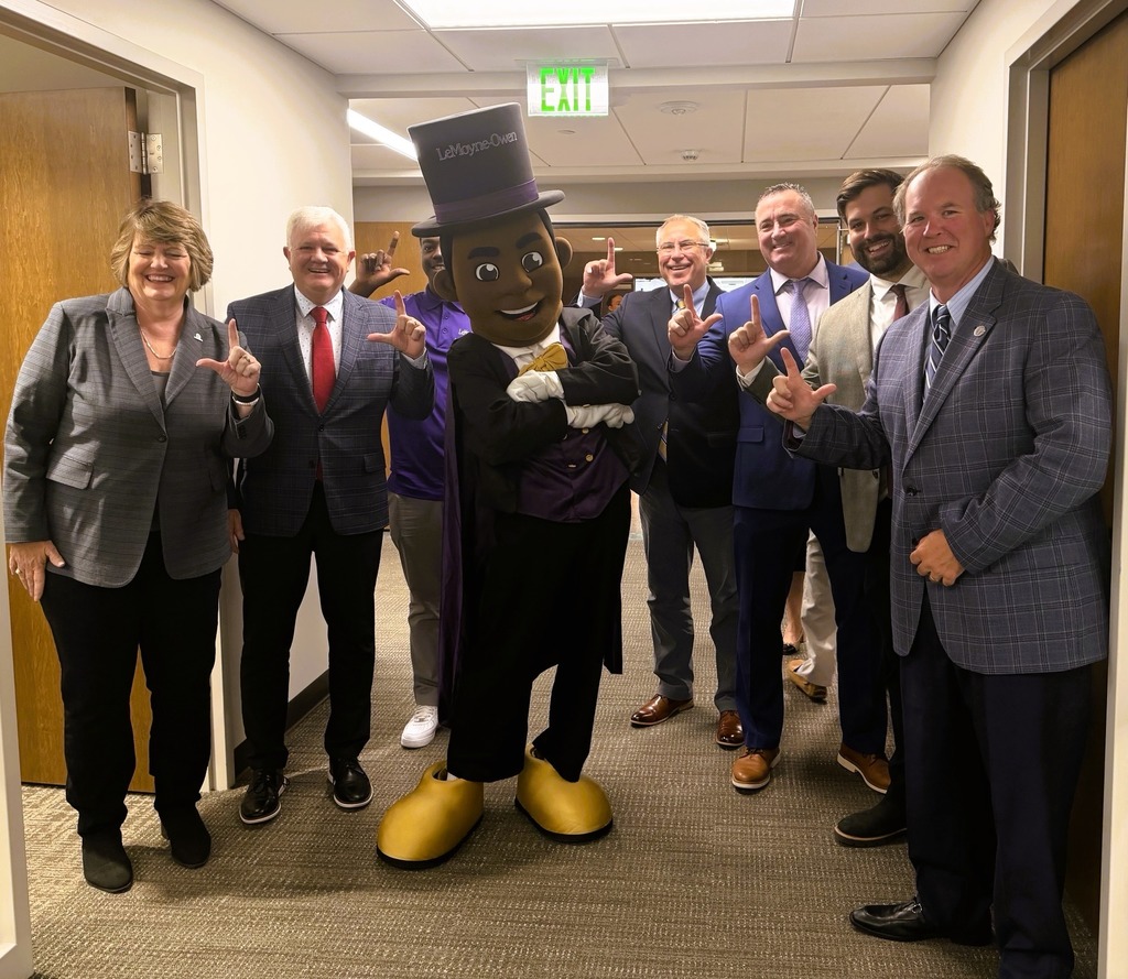 Group of political professionals posing in a hallway with the mascot from LeMoyne-Owen College, all making L shapes with their hands