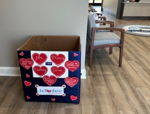 Photo of a Clothing Drive Box covered with red hearts and a LeBonheur sticker.  Box is sitting on a wood floor against a beige wall of an office lobby. 